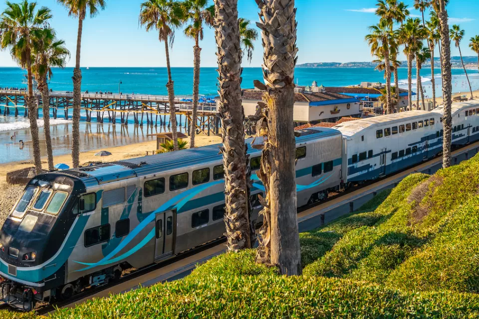 Silver Metrolink commuter train on tracks next to beach in San Clemente, California with palm trees and wooden pier in background.